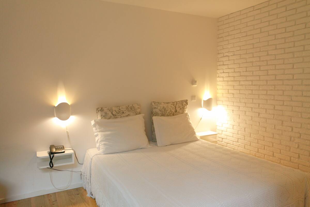 Un dormitorio con pared de ladrillo blanco, cama con ropa de cama blanca, dos lámparas de noche y un teléfono en la mesita.
