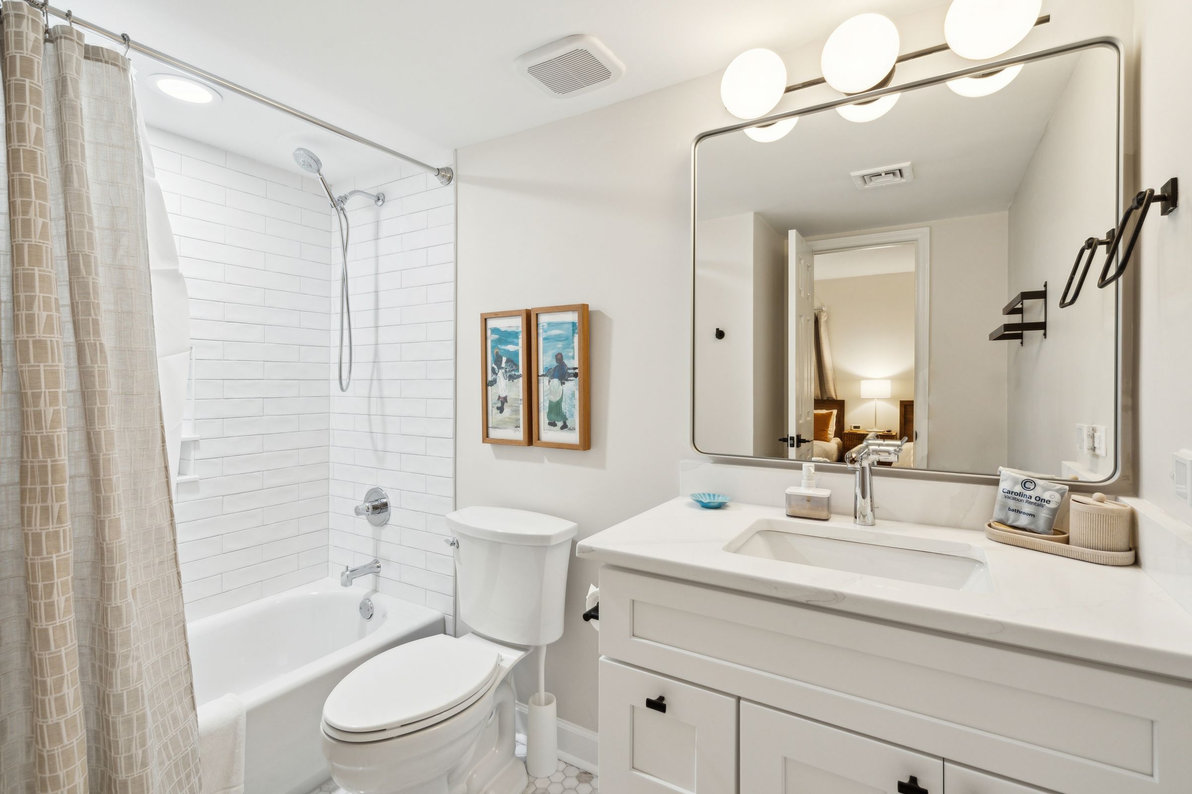 A modern bathroom with a bathtub, toilet, and sink. There are framed pictures on the wall and a mirror with lights above the sink.