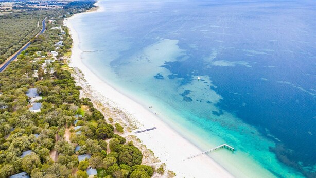 An aerial view of a coastal area with a long sandy beach, a road running parallel to the coast, and a pier extending into the water. - Siesta Park Holiday Resort (Busselton)