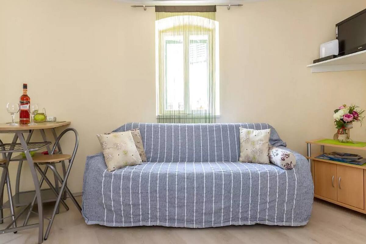 Una habitación pequeña y acogedora con un sofá a rayas, una mesita con una botella y vasos, un armario con un jarrón de flores y una ventana con cortinas.