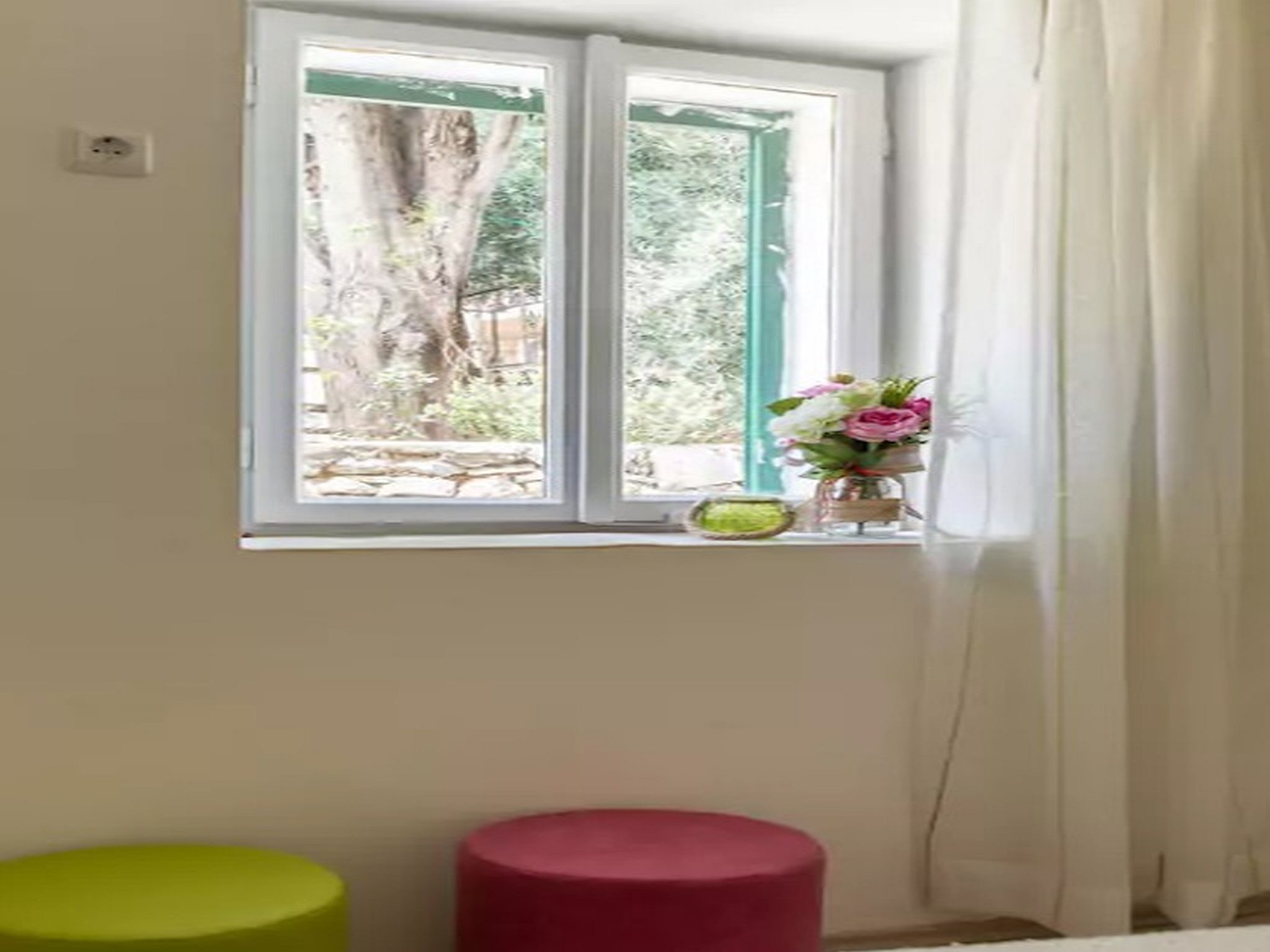 A room with a window, a green stool, a red stool, and a vase with flowers on the windowsill.