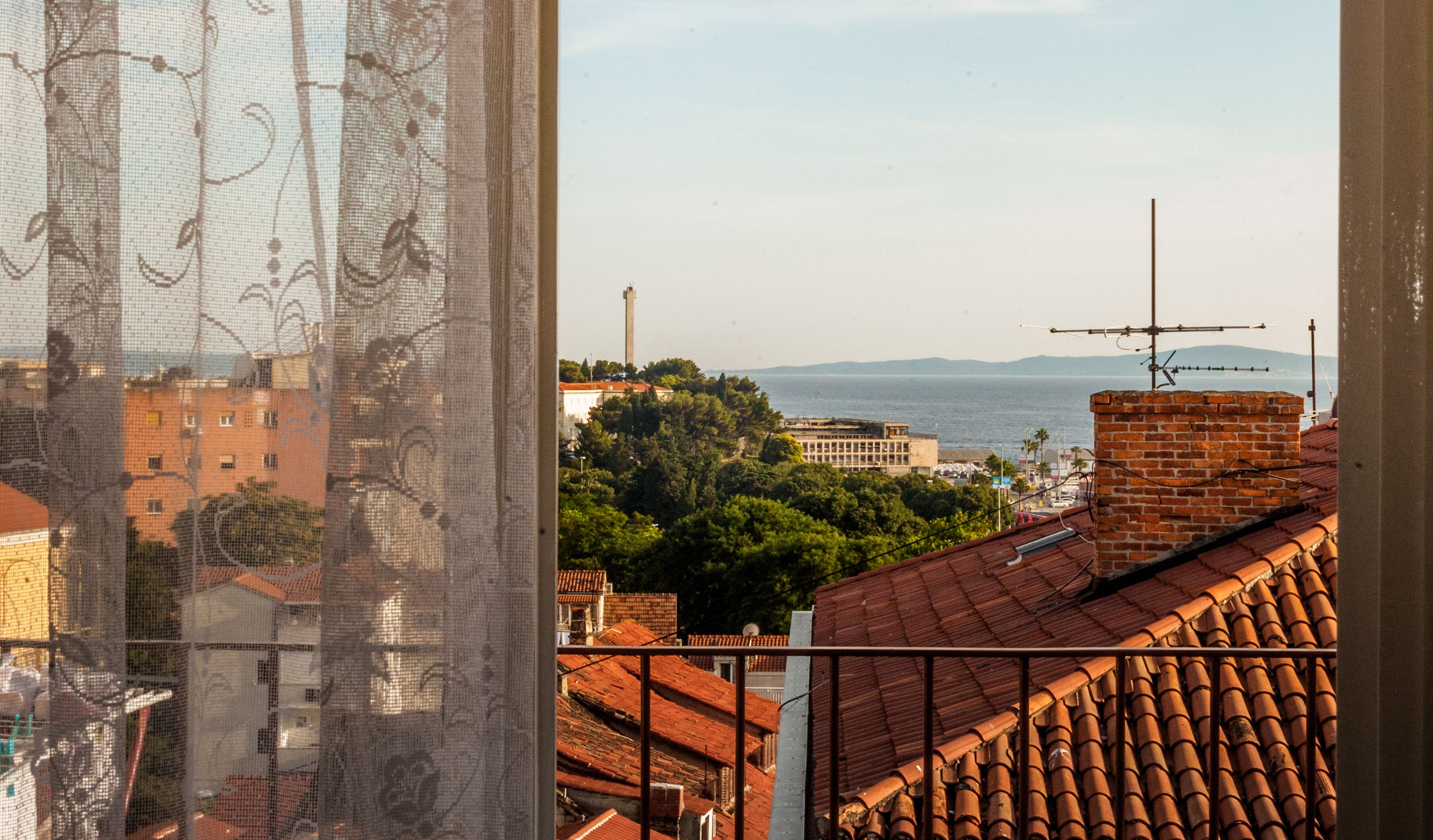 A view through a window with a mesh screen, featuring a coastal cityscape with buildings, a lighthouse, and the sea.