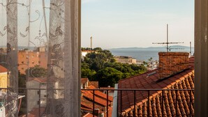 Vista a través de una ventana con mosquitera, que muestra un paisaje urbano costero con edificios, un faro y el mar.