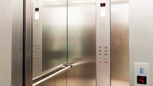 Interior de un ascensor con paredes de acero inoxidable, barandilla y paneles de control con botones de emergencia e indicadores de piso.