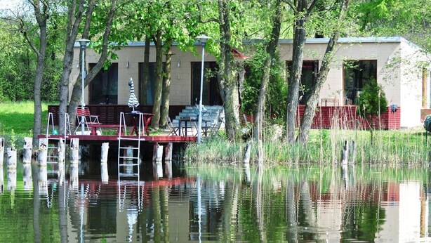 Een vakantiehuis aan het meer met een houten steiger, een overdekte zithoek en een gebouw met grote ramen.