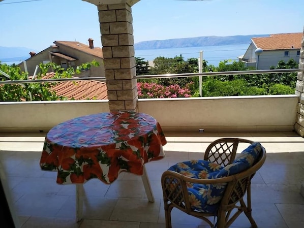 A balcony with a round table covered in a floral tablecloth, a wicker chair with a cushion, and a view of the sea and mountains in the background.