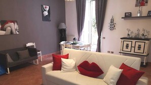 A living room with a white sofa, red and white cushions, a black floor lamp, a small table with a lamp, and a cabinet with decorative items. - Chic 'n Charme Trastevere Terrace (Roma)