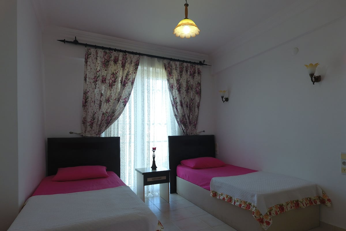 A room with two beds, each with a white bedspread and a floral border, pink pillows, and a small table with a vase between the beds.