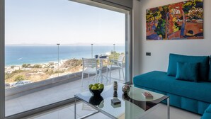 A modern living room with a large window overlooking a coastal view, a blue sofa, and a glass coffee table with decorative items. - Rhodes Skyline Suites (Rhodes)