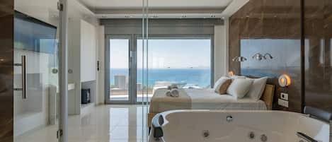 A modern hotel room with a large bed, a bathtub, a glass shower enclosure, and a view of the ocean.