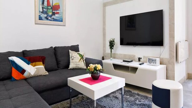 A modern living room with a sectional sofa, a flat-screen TV mounted on the wall, a coffee table with a vase of flowers, and a small side table with a radio. - Apartment Rita (Split)