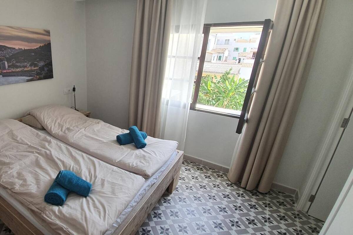 A room with two beds, a small bedside table, a lamp, a window with curtains, and a cityscape painting on the wall.