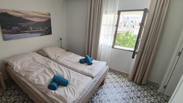 A room with two beds, a small bedside table, a lamp, a window with curtains, and a cityscape painting on the wall.