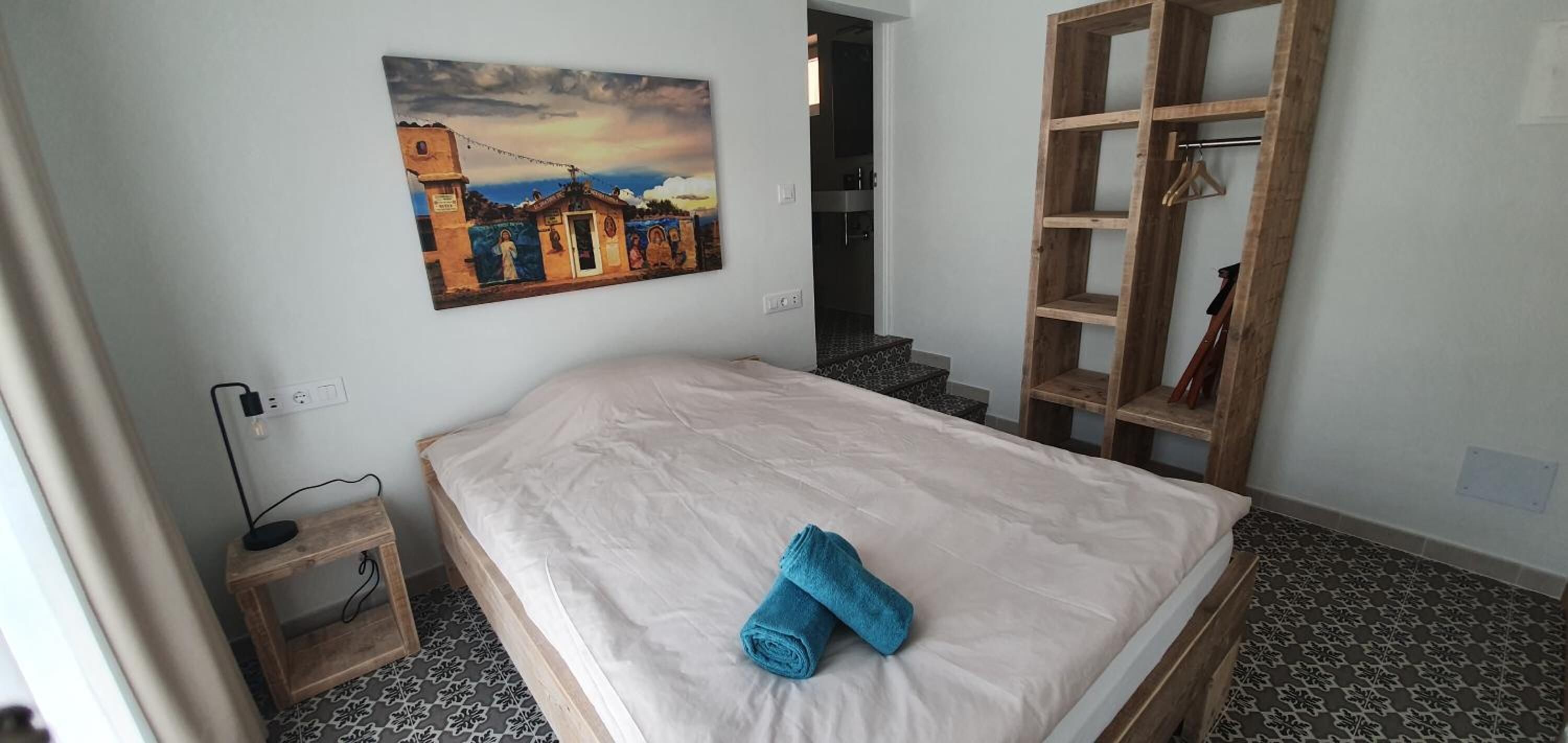 Ein Schlafzimmer mit einem Bett, einem Gemälde, einer Holzregal und einem durch eine offene Tür einsehbaren Badezimmer.