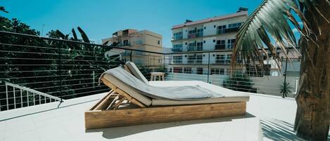 Ein Einzelbett aus Holz mit einer Matratze auf einem Balkon mit Blick auf Gebäude und eine Palme.