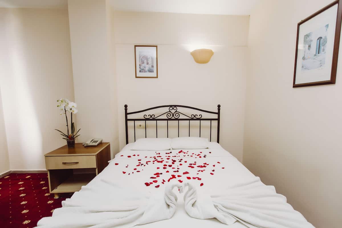 A neatly made bed with rose petals and a towel arranged in a heart shape, a bedside table with a phone, a framed picture on the wall, and a lamp.