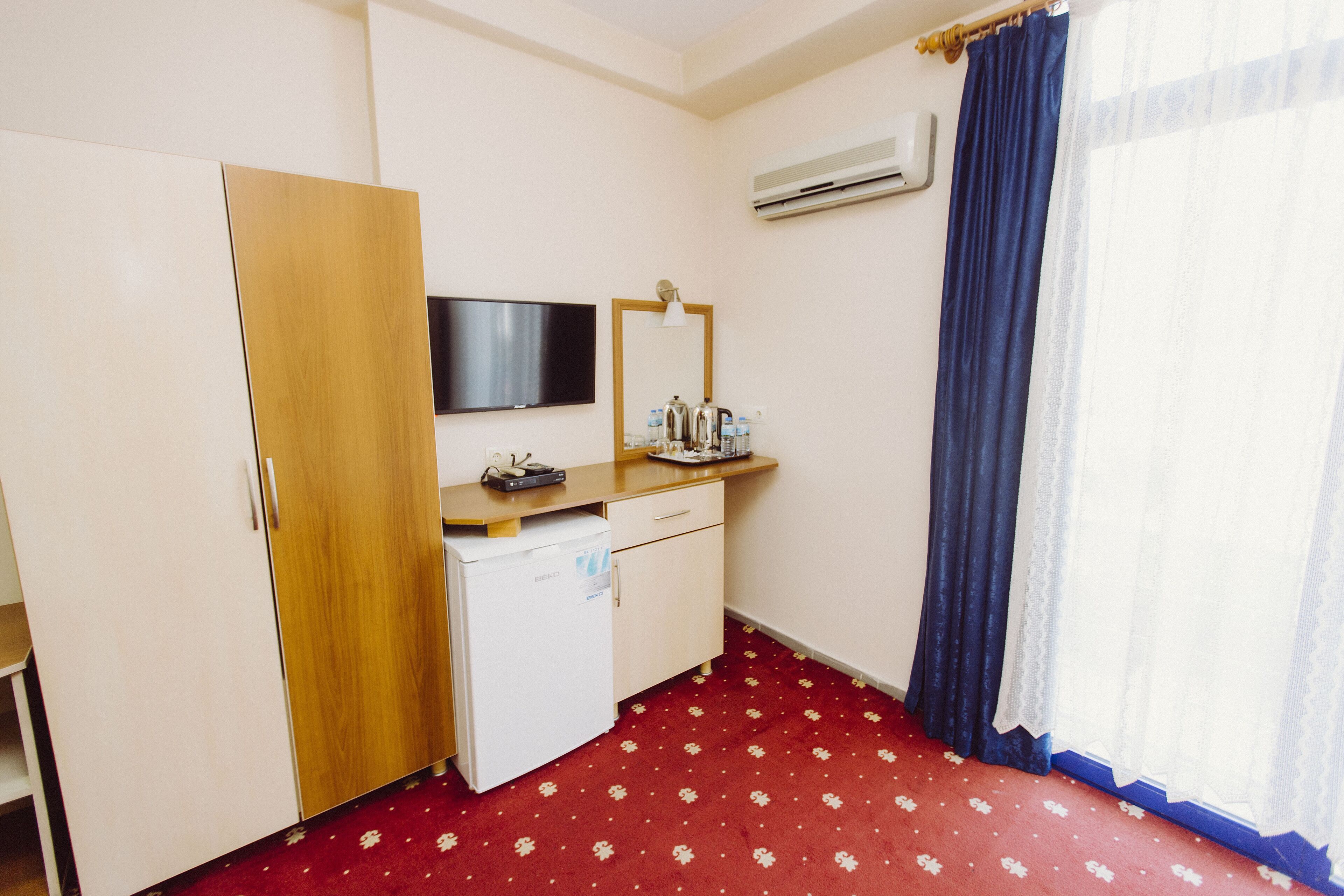 A hotel room with a wooden wardrobe, a small desk with a flat-screen TV, a mirror, and a red floral carpet.