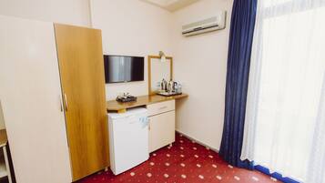 A hotel room with a wooden wardrobe, a small desk with a flat-screen TV, a mirror, and a red floral carpet.