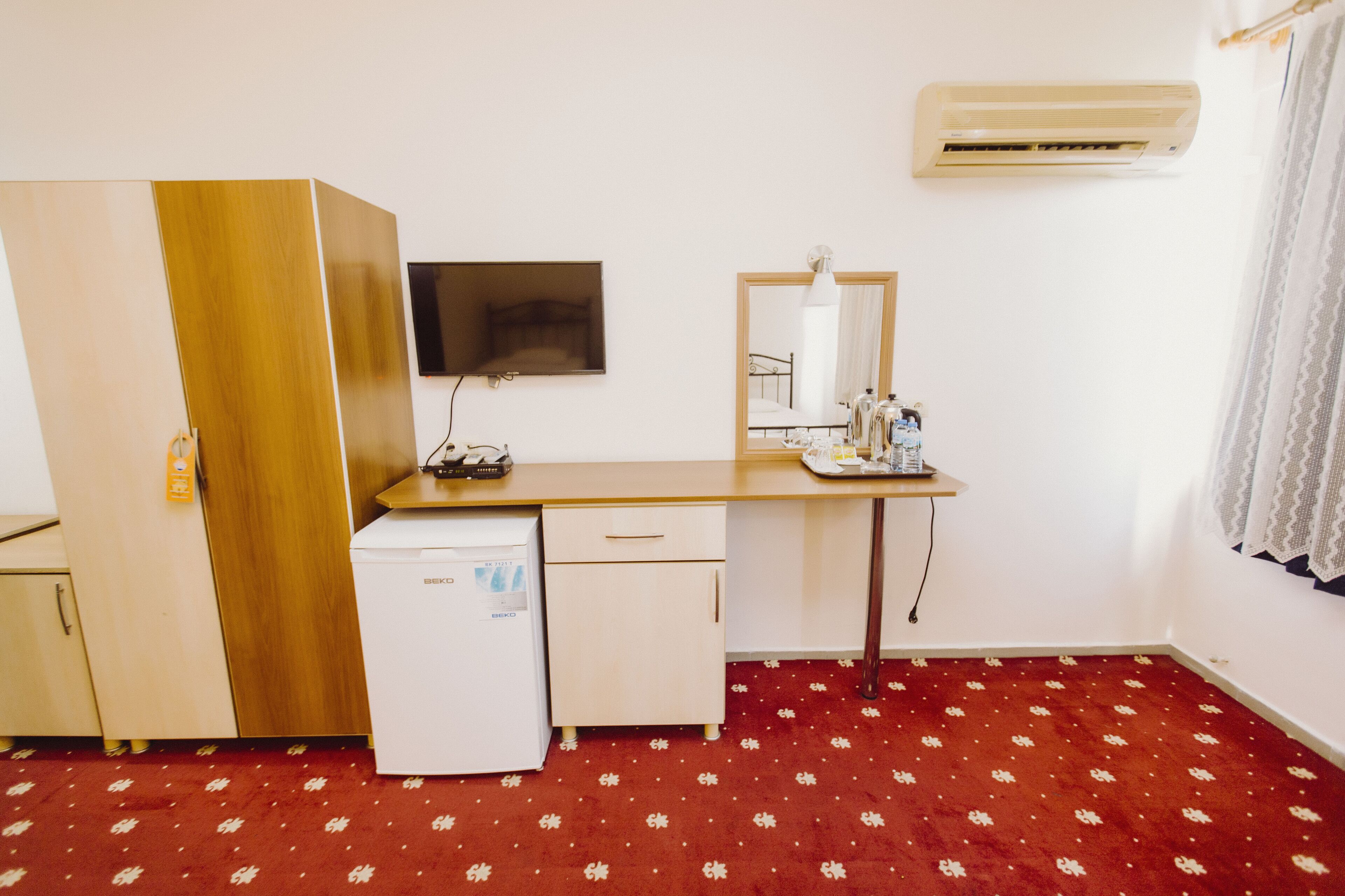 A compact room with a kitchenette, a small refrigerator, a flat-screen TV, a mirror, and a red patterned carpet.
