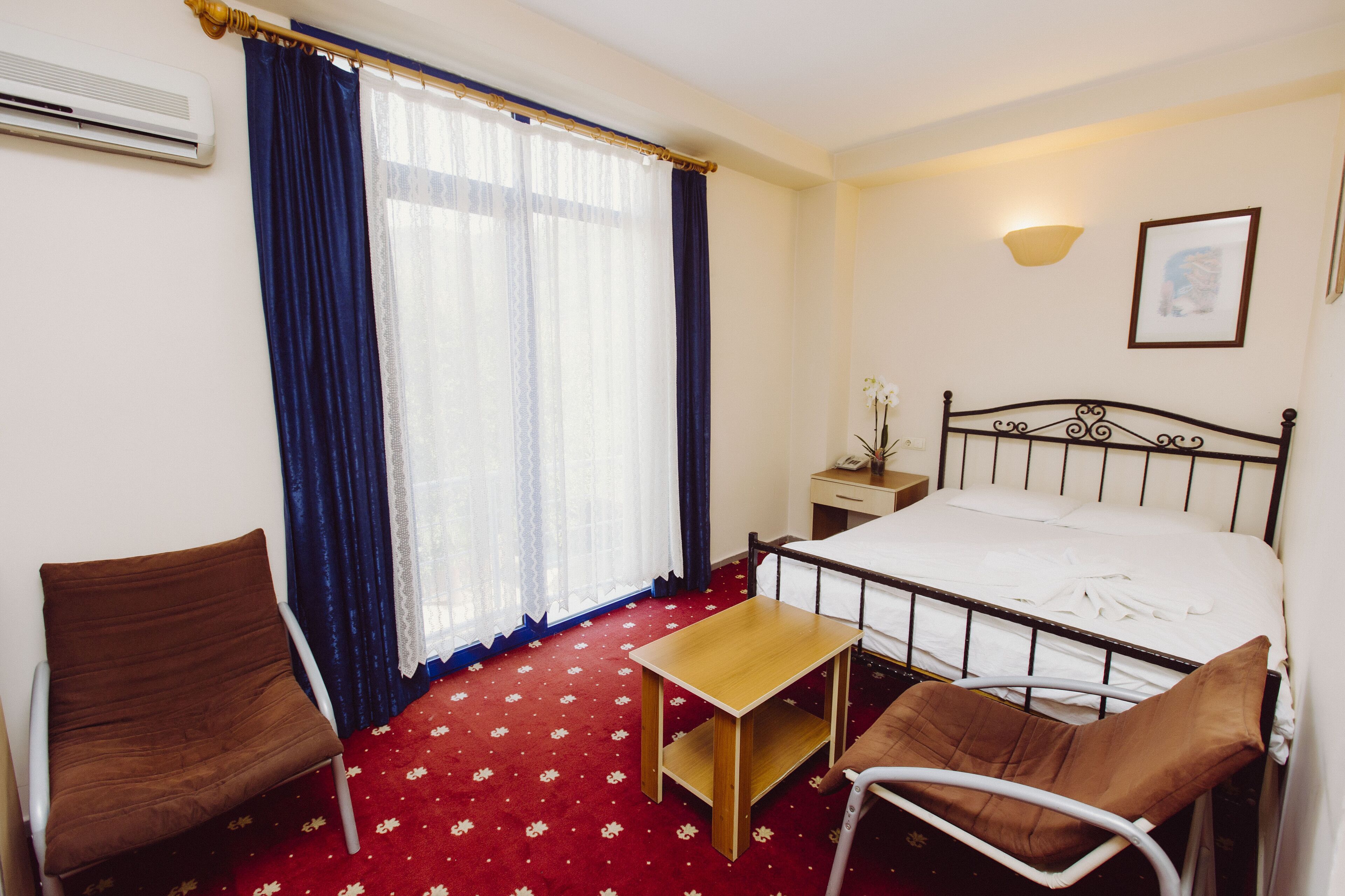 A hotel room with a bed, a chair, a small table, a nightstand, and a window with curtains.