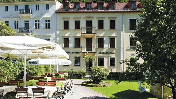 An outdoor seating area with tables and chairs, a white umbrella, and a building with multiple windows and balconies in the background. - Kurparkstübl Bad Schandau (Bad Schandau)