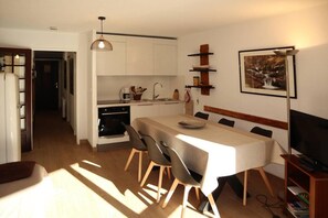 A compact kitchen with white cabinets, a dining table with chairs, a TV, and a bookshelf.