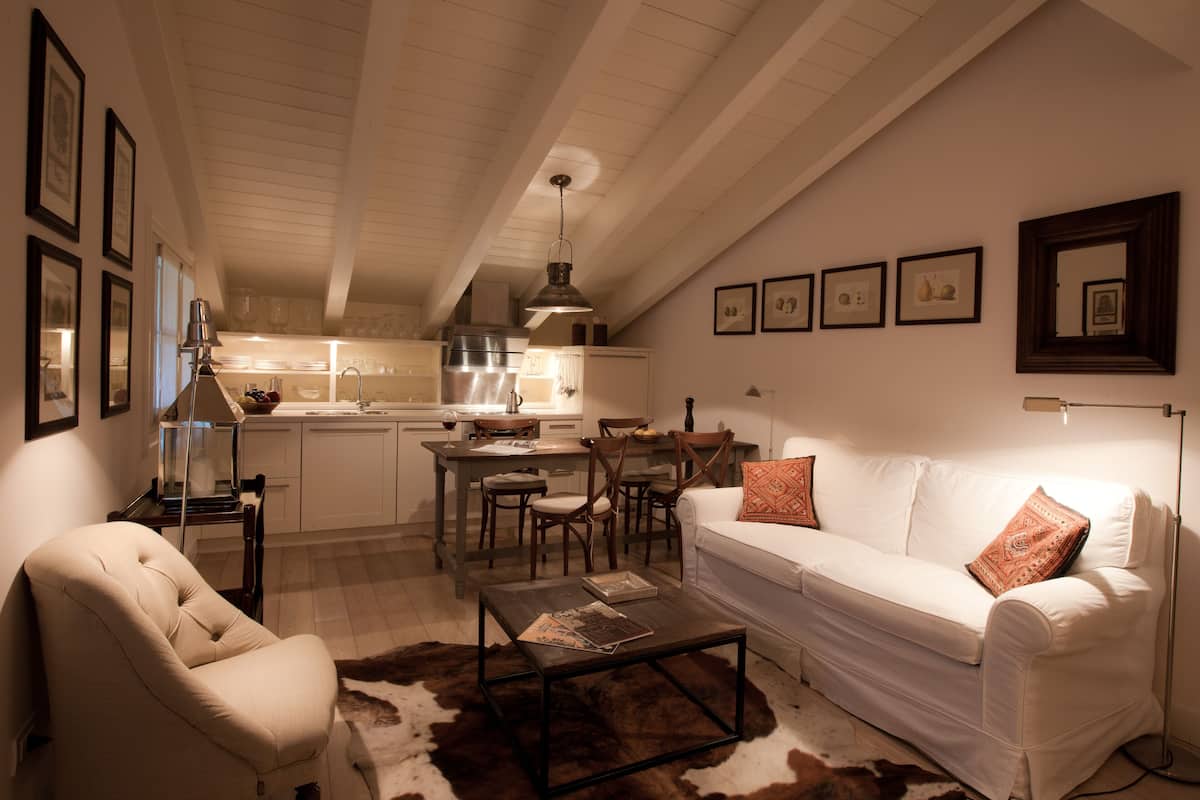 Una sala de estar acogedora con un sofá blanco, una mesa de centro y un tapiz de cuero de vaca. Se aprecia una cocina al fondo con armarios blancos y un comedor con sillas de madera.