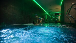 A dimly lit indoor swimming pool with green lighting, a person performing a flip, and a visible gym area in the background. - Les Fleurs Bleues chalets (Livigno)