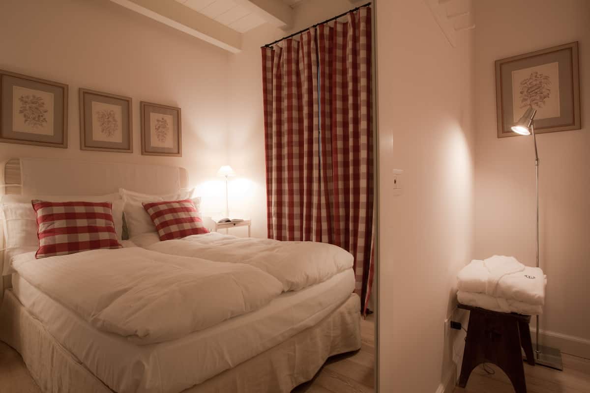 Un dormitorio con una cama grande, una mesita de noche con lámpara, una cortina a cuadros blancos y rojos, y cuadros en la pared.