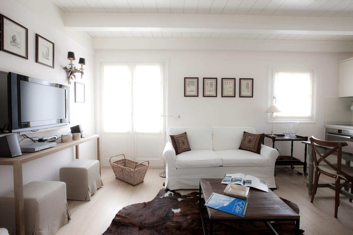 Una sala de estar moderna con un sofá blanco, una mesa de centro de madera y un televisor sobre un mueble de madera. Hay cuadros enmarcados en la pared y una ventana con persianas.