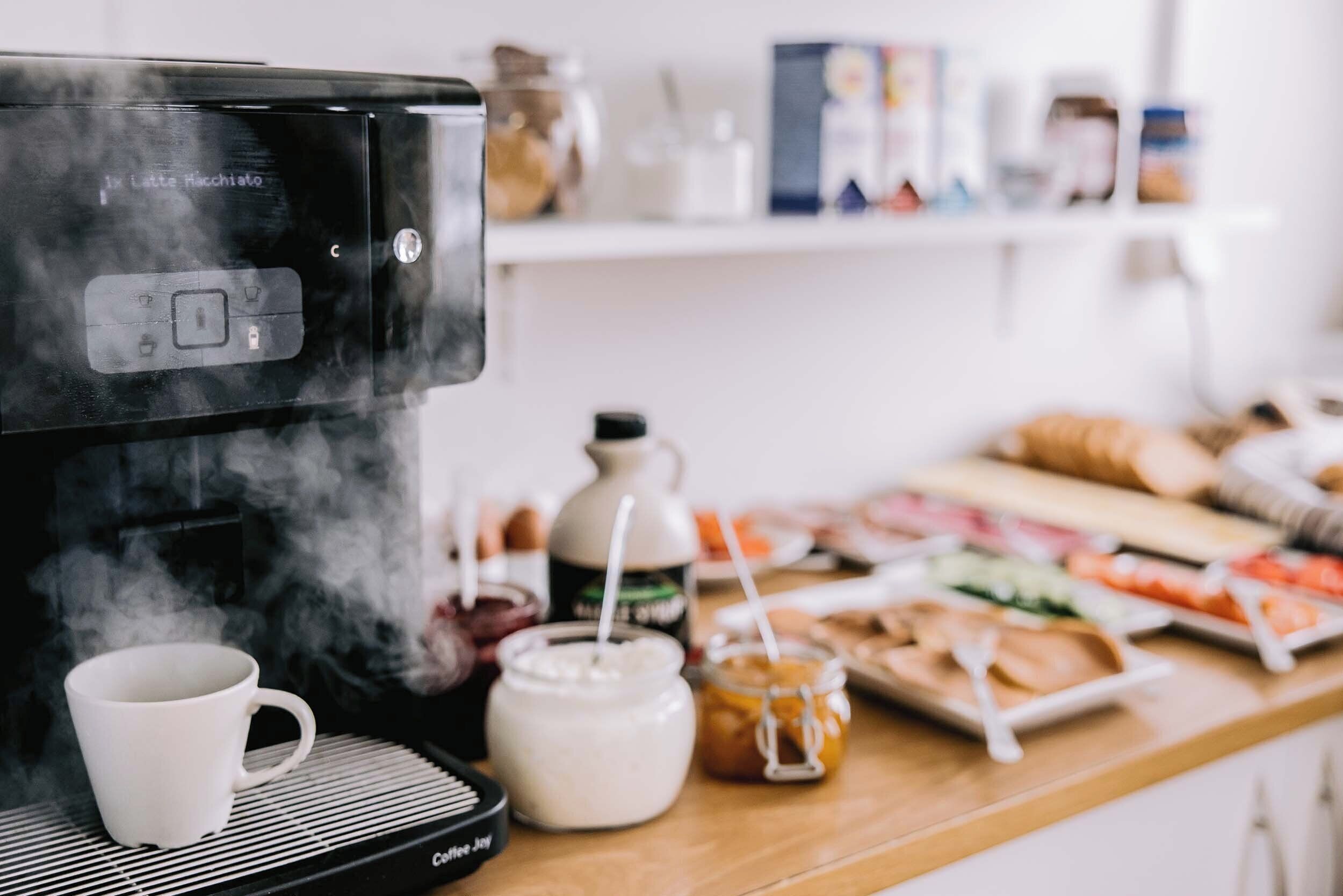 Una cafetera con vapor, una taza blanca y una bandeja de desayuno sobre una mesa de madera.