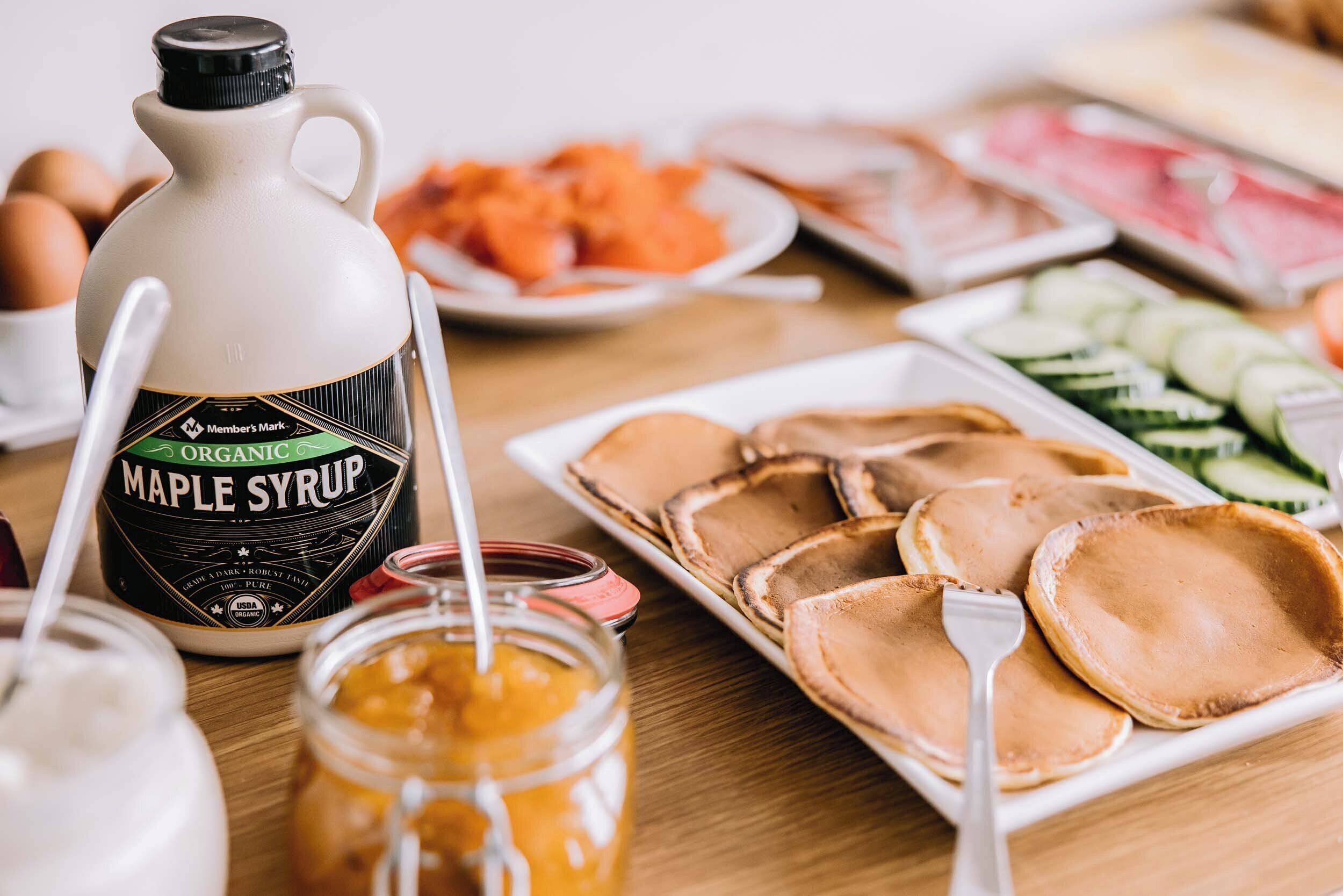 A breakfast buffet with various food items including pancakes, eggs, and a bottle of maple syrup.