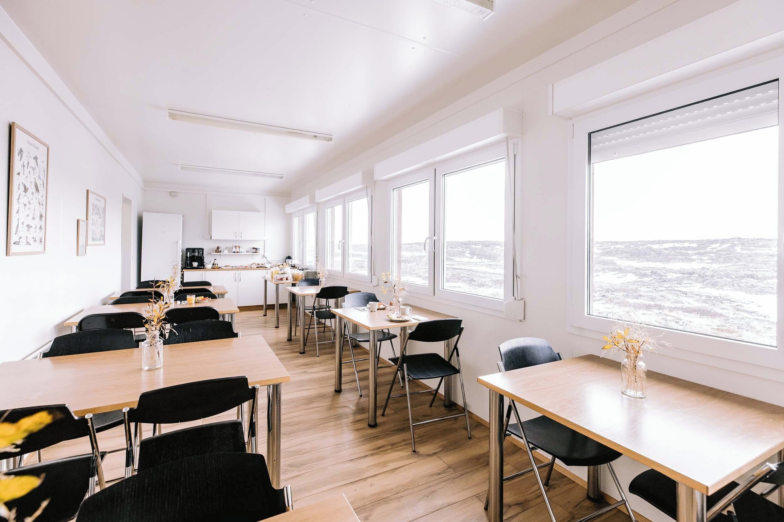 A modern, well-lit dining area with multiple tables and chairs, large windows offering a view of the sea, and a kitchen in the background.