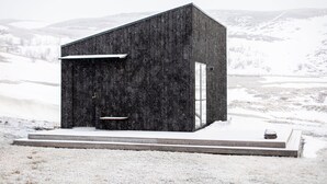Una cabaña moderna de madera negra con techo plano, ubicada en una colina nevada con vistas a montañas distantes.