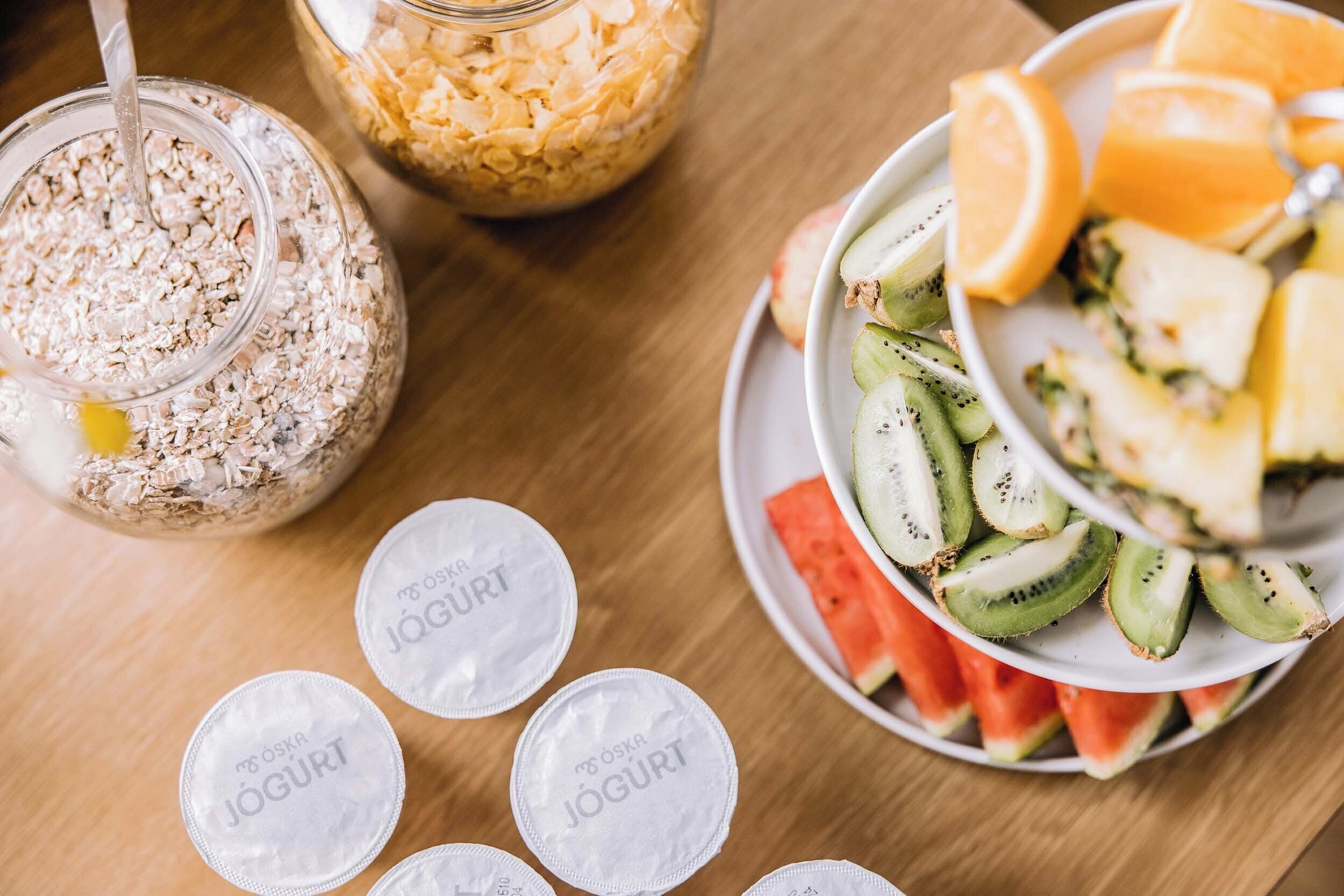 Una mesa con un tazón de frutas en rodajas, un plato con vasitos de yogur y un tarro de avena.