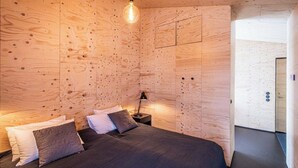 Un dormitorio con paneles de madera en la pared, una cama con ropa de cama oscura, una lámpara de noche y una bombilla colgando del techo.