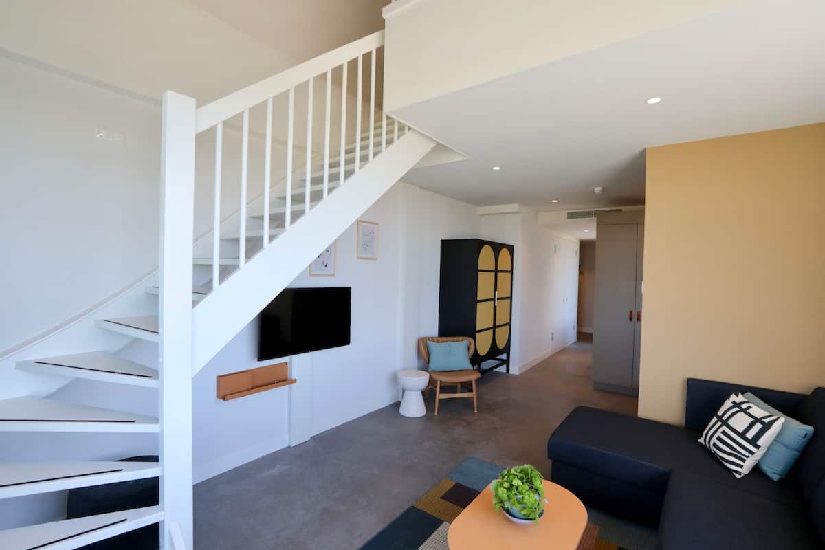 Una sala de estar moderna con una escalera, un televisor de pantalla plana montado en la pared, un sofá con cojines, una pequeña mesa redonda con una planta y un separador decorativo.