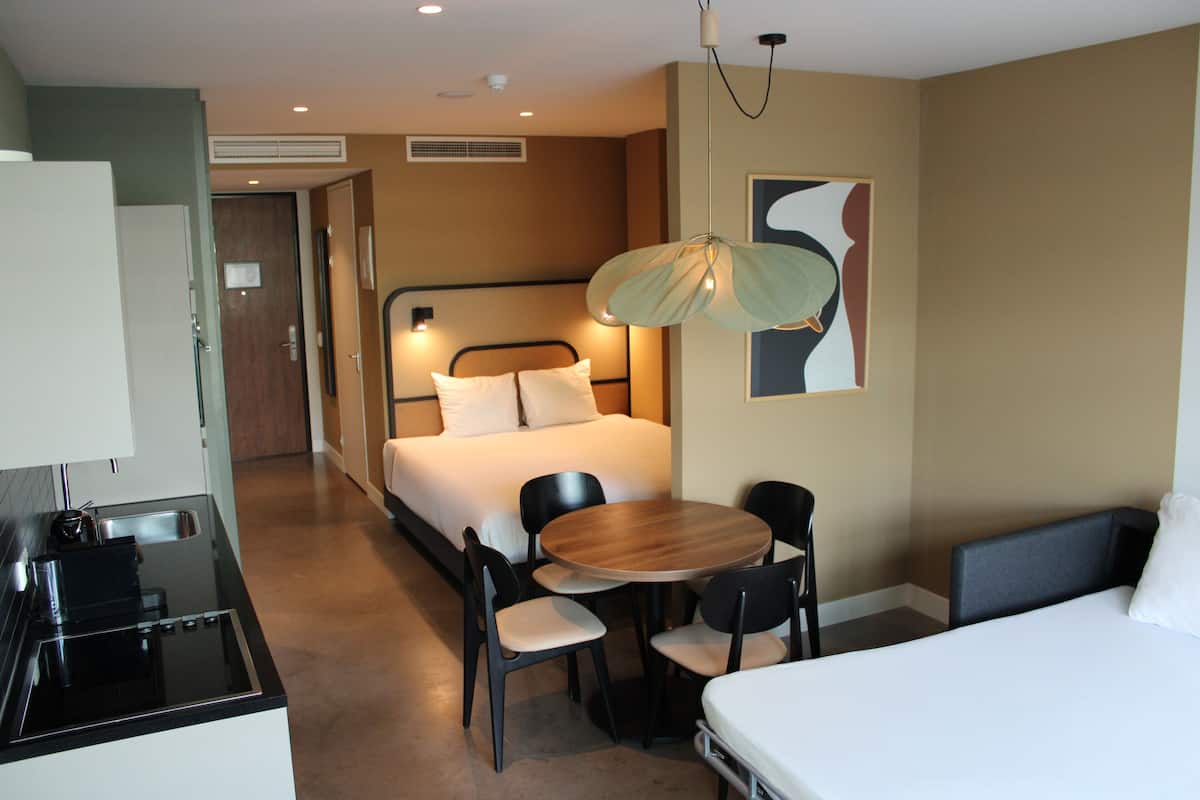 Habitación de hotel con cama, escritorio con silla, una mesa redonda con dos sillas y una lámpara colgante.