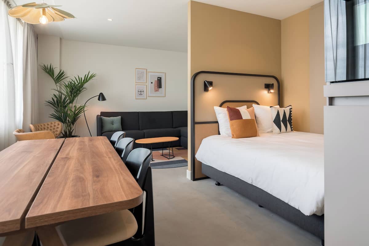 Una habitación de hotel moderna con una mesa de comedor de madera, un sofá, una cama con cabecera y una planta en maceta.