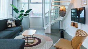 Ein modernes Wohnzimmer mit einer Couch, einem Couchtisch, einem Sessel, einer Zimmerpflanze, einer Stehlampe und einem an der Wand montierten Fernseher.
