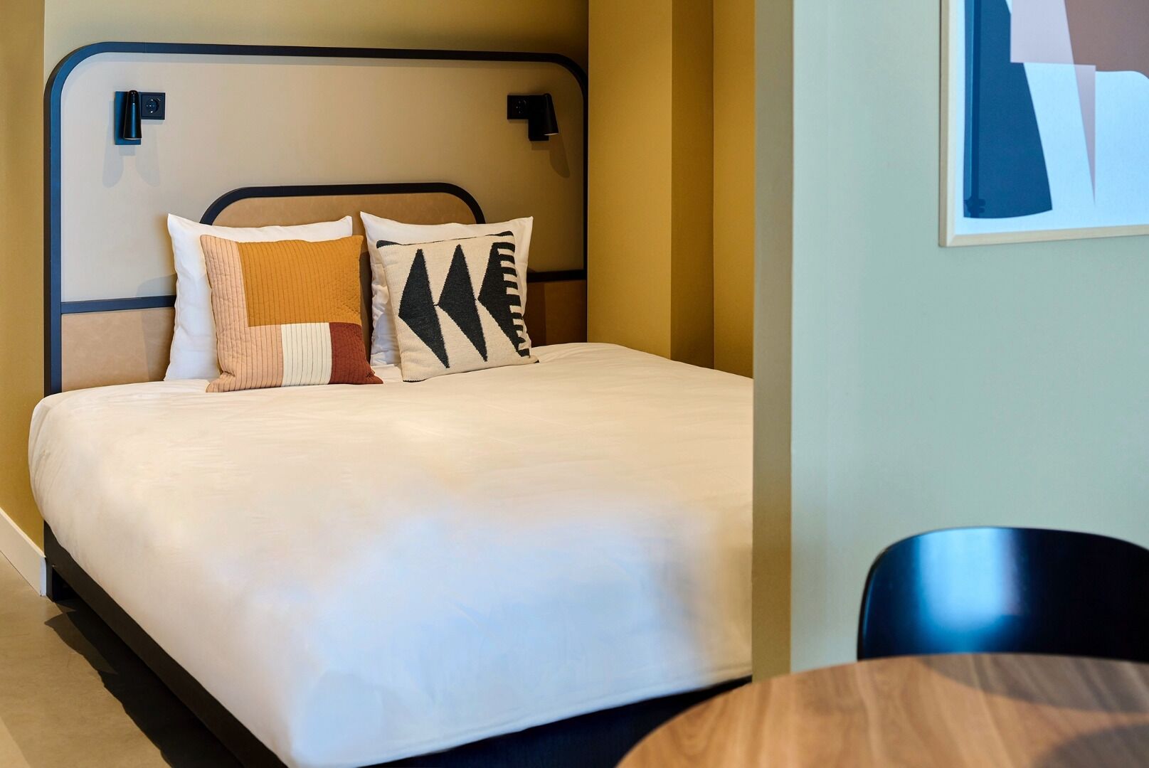 Een net opgemaakt bed met een witte deken, twee kussens met geometrische patronen en een houten nachtkastje met een lamp.
