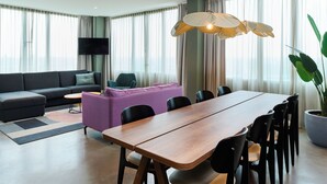 Ein modernes Wohnzimmer mit einer Couch, einem Esstisch mit Stühlen, einem Fernseher und einem großen Fenster mit Vorhängen.