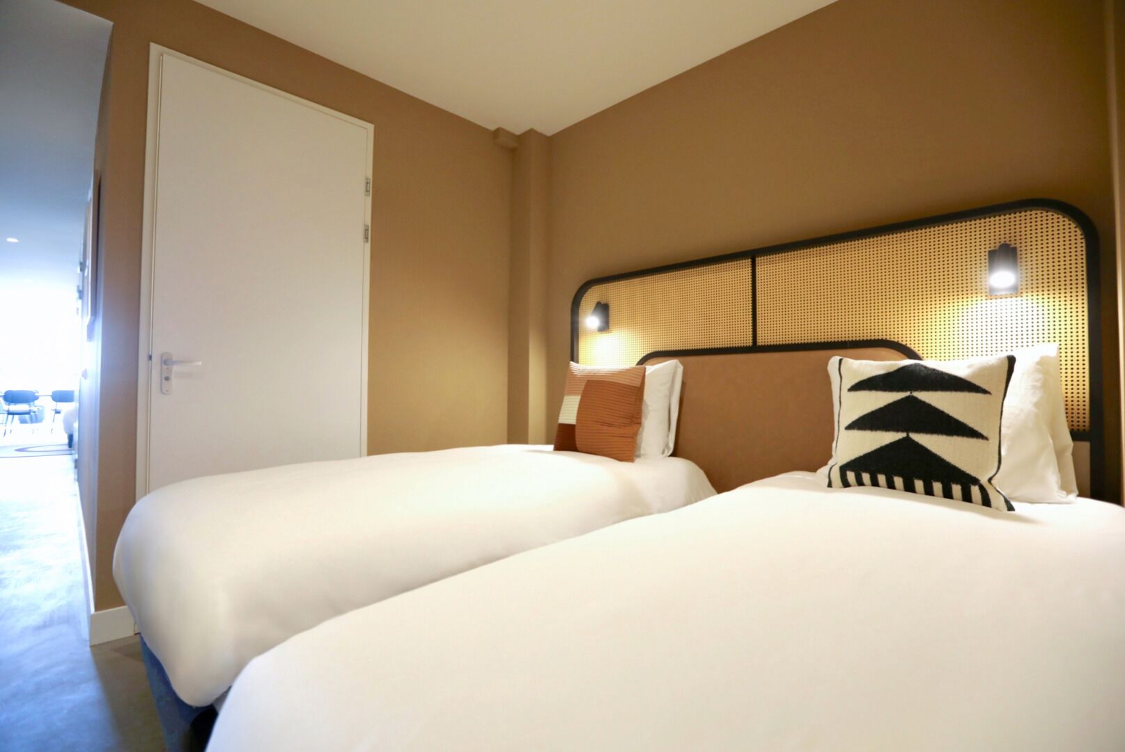 Habitación de hotel con dos camas, una puerta y un cabecero trenzado.