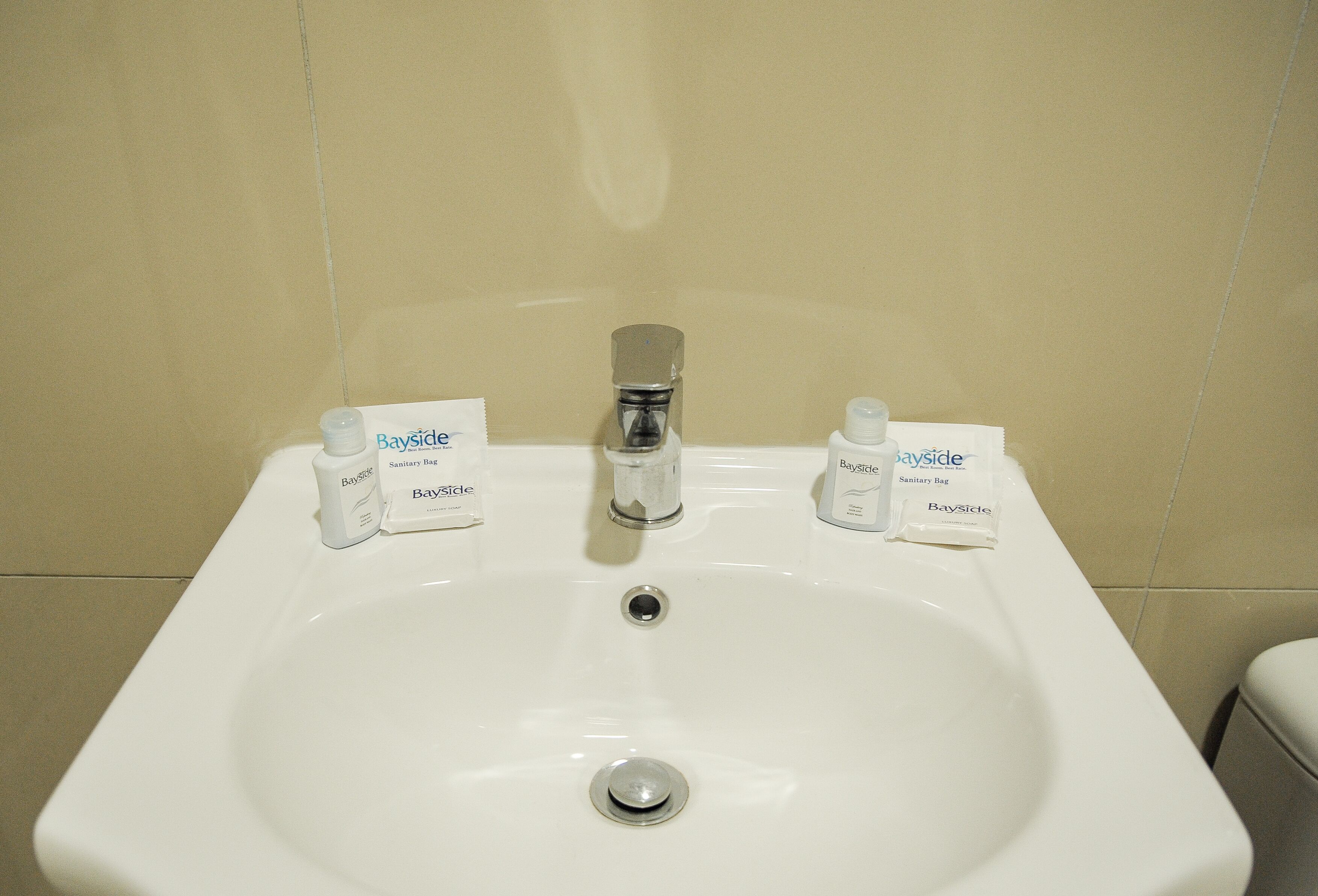 Un lavabo bianco con rubinetto cromato, due sacchetti igienici con l'etichetta 'Bayside' e pareti rivestite di piastrelle color beige.