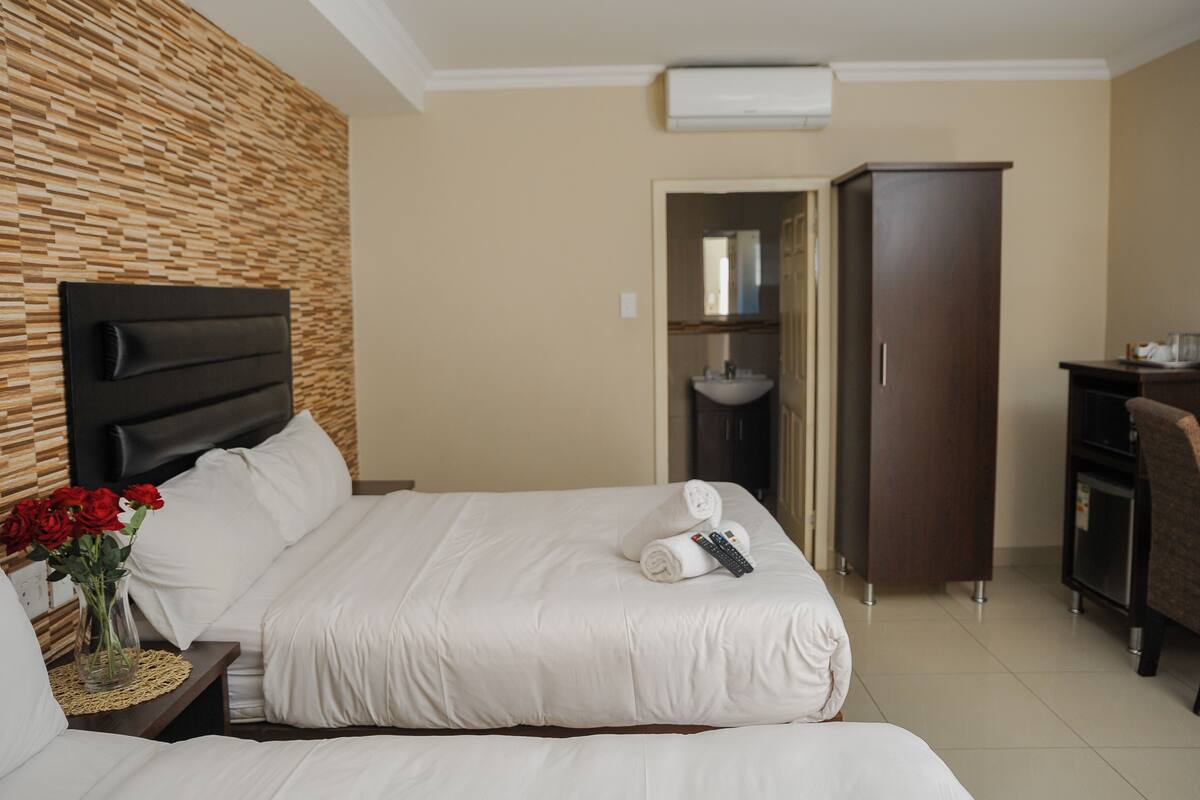 A hotel room with a bed, a nightstand with a vase of red roses, a wooden headboard, a wardrobe, and a desk with a chair.