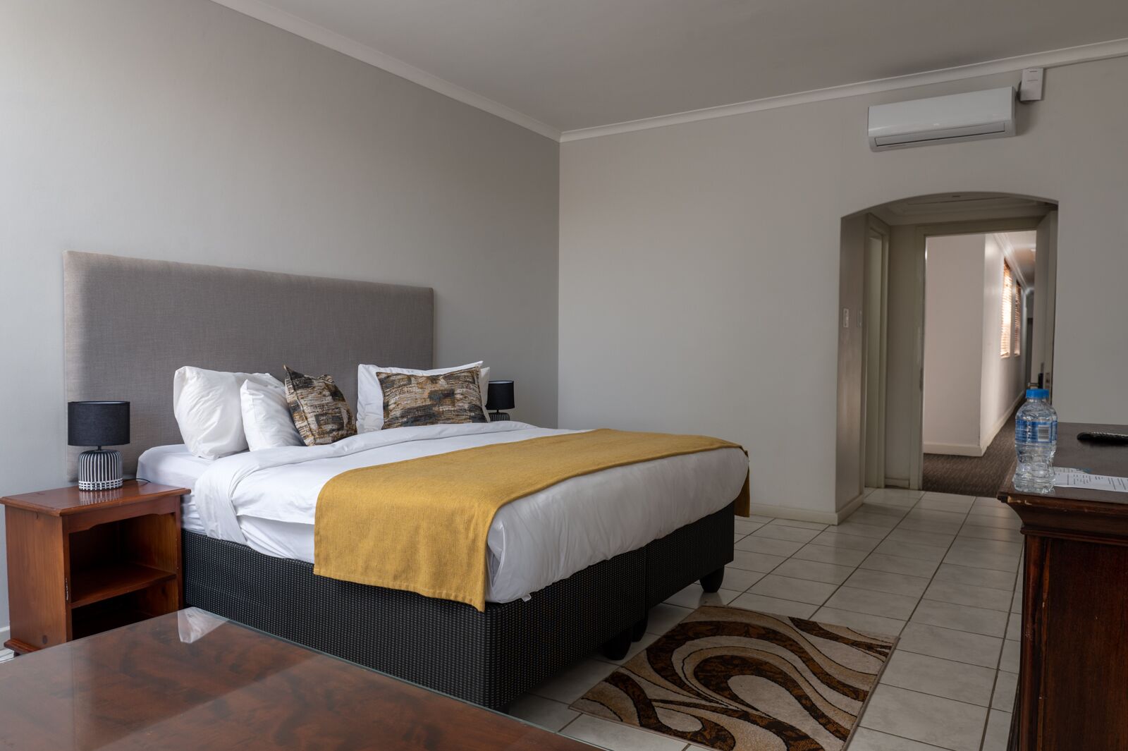 A hotel room with a bed, bedside tables, a desk, and an air conditioning unit.