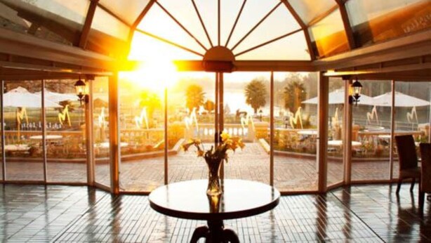 A glass-enclosed patio with a round table and chairs, a vase with flowers, and a view of the outdoors. - Riviera On Vaal Resort (Vereeniging)