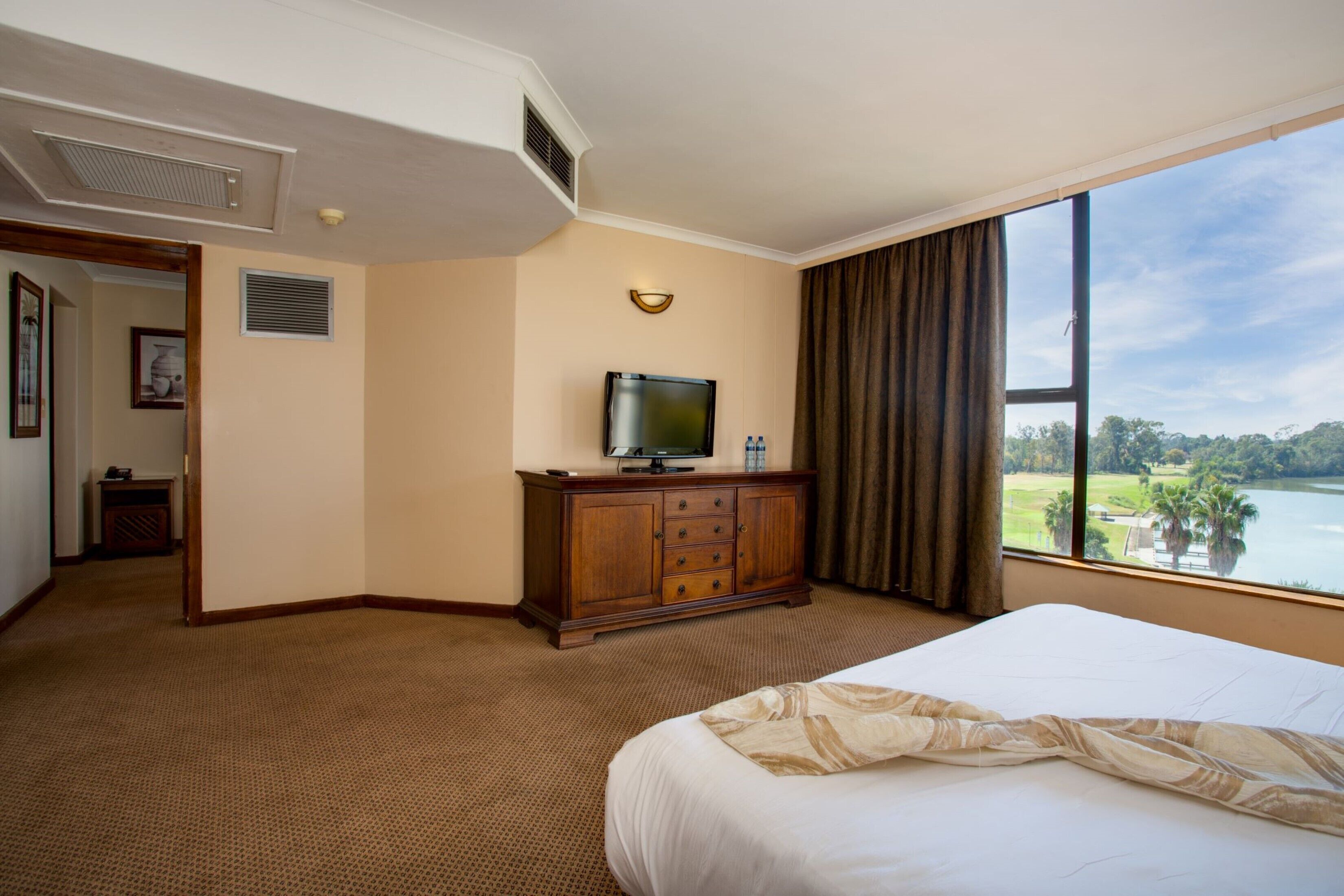 Een hotelkamer met een groot bed, een televisie op een houten kast, uitzicht op een meer, en een deur naar een andere kamer.