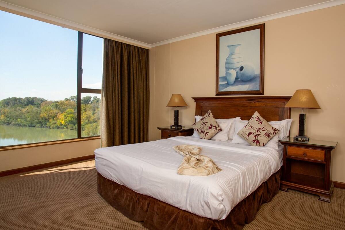 A hotel room with a large bed, two bedside tables, a nightstand, a painting above the bed, and a view of a lake through a large window.
