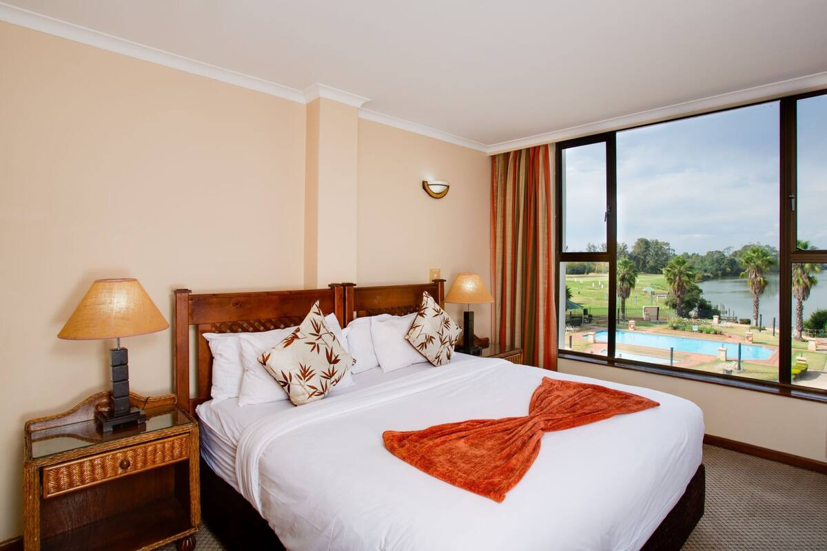 A hotel room with a large bed, two bedside lamps, a view of a pool and landscape through large windows.